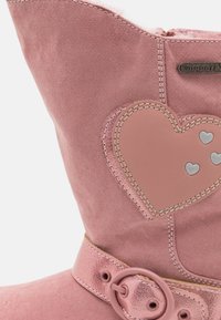 Světle růžová semišová bota se širokým chlopněm, zdobená nášivkou ve tvaru srdce, kovovými doplňky a zaoblenou sponou. Viditelný měkký vnitřek.