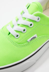 Kirkkaita vihreitä Vans-tossuja, joissa valkoiset nauhat ja pohja. Lähellä otettu kuva, joka näyttää tekstiilin rakenteen, silmukat ja Vans-logotagin sivussa.