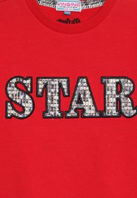 Sudadera roja con la palabra "STAR" en letras grandes y texturizadas, con un patrón tejido multicolor y contorno negro. Etiqueta del fabricante visible.