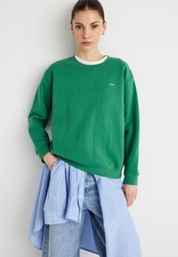 Sweat-shirt en coton vert à coupe décontractée avec des poignets côtelés, superposé sur une chemise rayée bleu et blanc, associé à un jean clair.