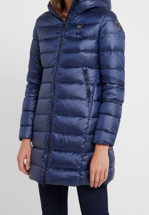 Kobieta ubrana w granatową, pikowaną kurtkę puchową z długim zamkiem, kapturem, zapinaną na zamek boczną kieszeń oraz naszywki z logo na piersi i rękawie.