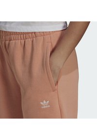 Ljust rosa sweatpants med elastisk midja, sidofickor och en vit Adidas-logotyp nära vänster ficka. Mjuk tygtextur.