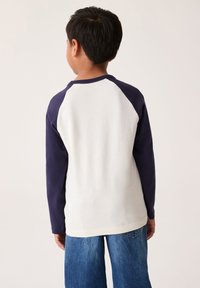 Majica z dolgimi rokavi z belim telesom in mornarsko modrimi raglan rokavi, z okroglim izrezom in ravnim spodnjim robom. Mehka tkanina.