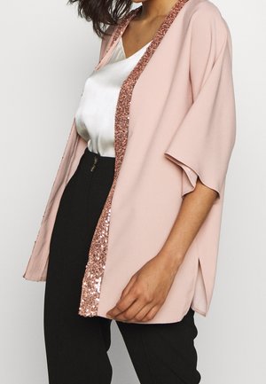 Kobieta ubrana w jasnoróżowe kimono z otwartym przodem i wykończeniem z cekinów, białą satynową bluzkę oraz czarne spodnie z wysokim stanem na jednolitym tle.