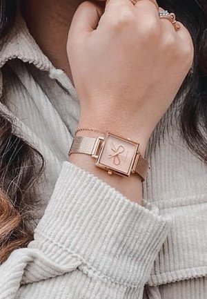 Poignet portant une montre carrée en or rose avec un motif de nœud sur le cadran, portée sur un poignet de pull côtelé beige clair.