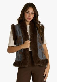 Gilet di jeans con finiture in pelliccia sintetica marrone, decorato con ricami floreali in marrone. Design senza maniche e tasche frontali. Abbinato a una maglia neutra.