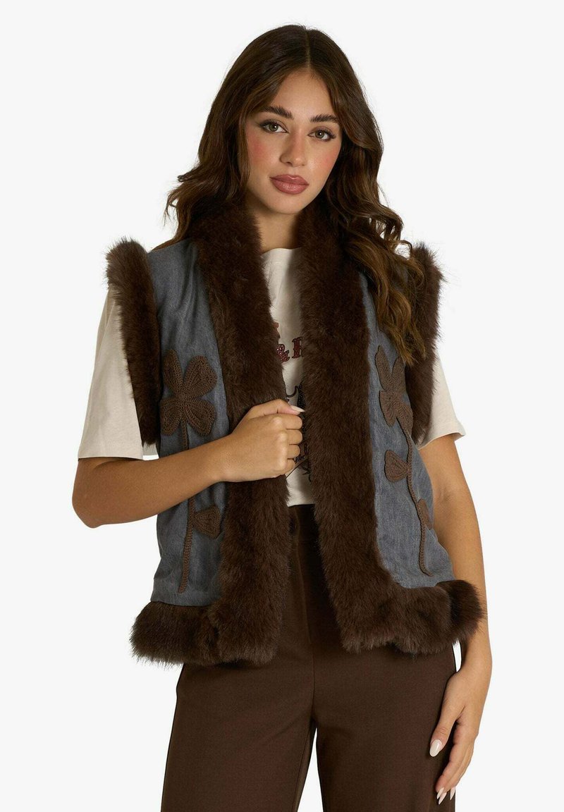 Gilet di jeans con finiture in pelliccia sintetica marrone, decorato con ricami floreali in marrone. Design senza maniche e tasche frontali. Abbinato a una maglia neutra.
