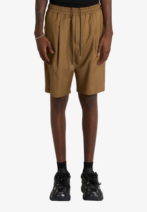 Persona che indossa pantaloncini marroni larghi con vita elastica e coulisse nere, sneakers nere e calzini neri su sfondo bianco.