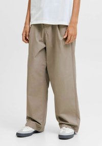 Bambino in pantaloni beige larghi, sneakers bianche con suole grigie e una maglietta bianca su uno sfondo chiaro e uniforme.