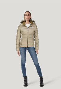 Giacca a vento trapuntata beige con chiusura zip e cappuccio, caratterizzata da un rivestimento interno verde a contrasto e un piccolo logo sul petto.