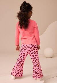 Top rosa de manga larga con costillas y puños con borde ondulado, combinado con pantalones acampanados rosa con estampado de leopardo; modelo de espaldas, calzando zapatillas blancas.