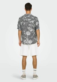 Kurzarmhemd in Grau mit floralem Muster, kombiniert mit weißen Shorts. Das Material scheint leicht zu sein; es hat einen lockeren Schnitt und einen Kragen.