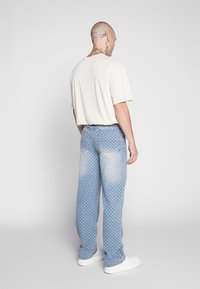 A person stands with their back facing the camera, wearing a loose beige t-shirt and patterned light blue jeans, and white sneakers.