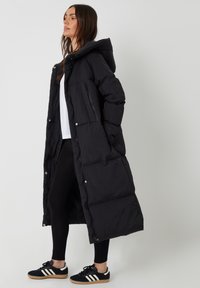 Zwarte lange pufferjas met een capuchon, voorzien van een doorgestikte stijl, twee voorkruiszakken en drukknoopsluitingen. Draagt over zwarte legging en sneakers.