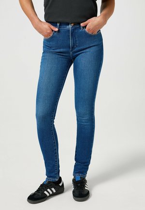 Jeans ajustados azules de cintura alta, con bolsillos frontales y detalles de costura. Llevados con zapatillas deportivas negras.