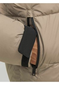 Primo piano di una giacca puffer beige con un cinturino in velcro nero e tasca con zip, che mette in evidenza un tessuto morbido e strutturato.