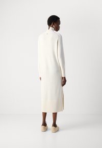 Robe longue, côtelée, de couleur crème avec un col haut et des fentes sur les côtés. La texture semble douce et lisse. Le modèle porte des chaussures beige.
