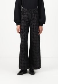 Schwarze weite Hosen aus Stoff mit einem subtilen Muster, hoher Taille und Fronttaschen. Kombiniert mit einem enganliegenden schwarzen Oberteil und schwarzen Schuhen.