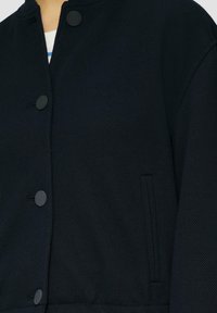 Veste bleu marine en tissu texturé, col rond et boutons noirs. Dotée d'une poche latérale et d'un design décontracté.