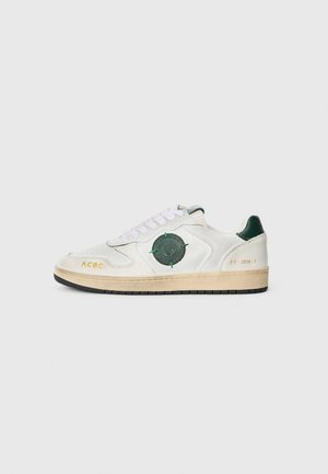 White sneaker with green accents, round patch, and gold text. Features lace-up design and textured rubber sole. Retro styling.