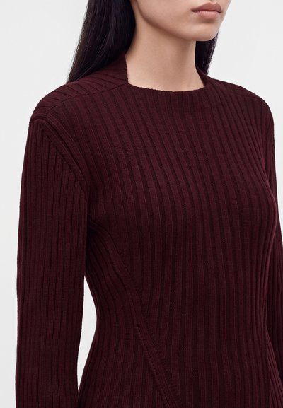 Femme portant un pull en maille côtelée bordeaux foncé avec manches longues et col rond, sur un fond uni.