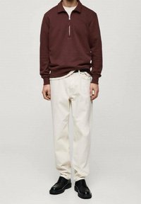 Pull à fermeture éclair bordeaux, pantalon beige clair, chaussures noires. Le pull a un col et une fermeture éclair métallique ; le pantalon est ample.