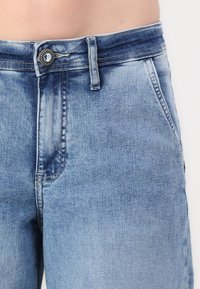 Svijetloplave traperice sa zatvaranjem na gumb, petljama za remen i prednjim džepovima. Tkanina pokazuje izblijedjeli izgled s blagim iznošenjem.