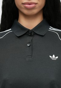 Μαύρο πόλο μπλουζάκι με γιακά, με τρεις λευκές ρίγες πάνω στους ώμους και ένα μικρό λευκό λογότυπο στο αριστερό στήθος.