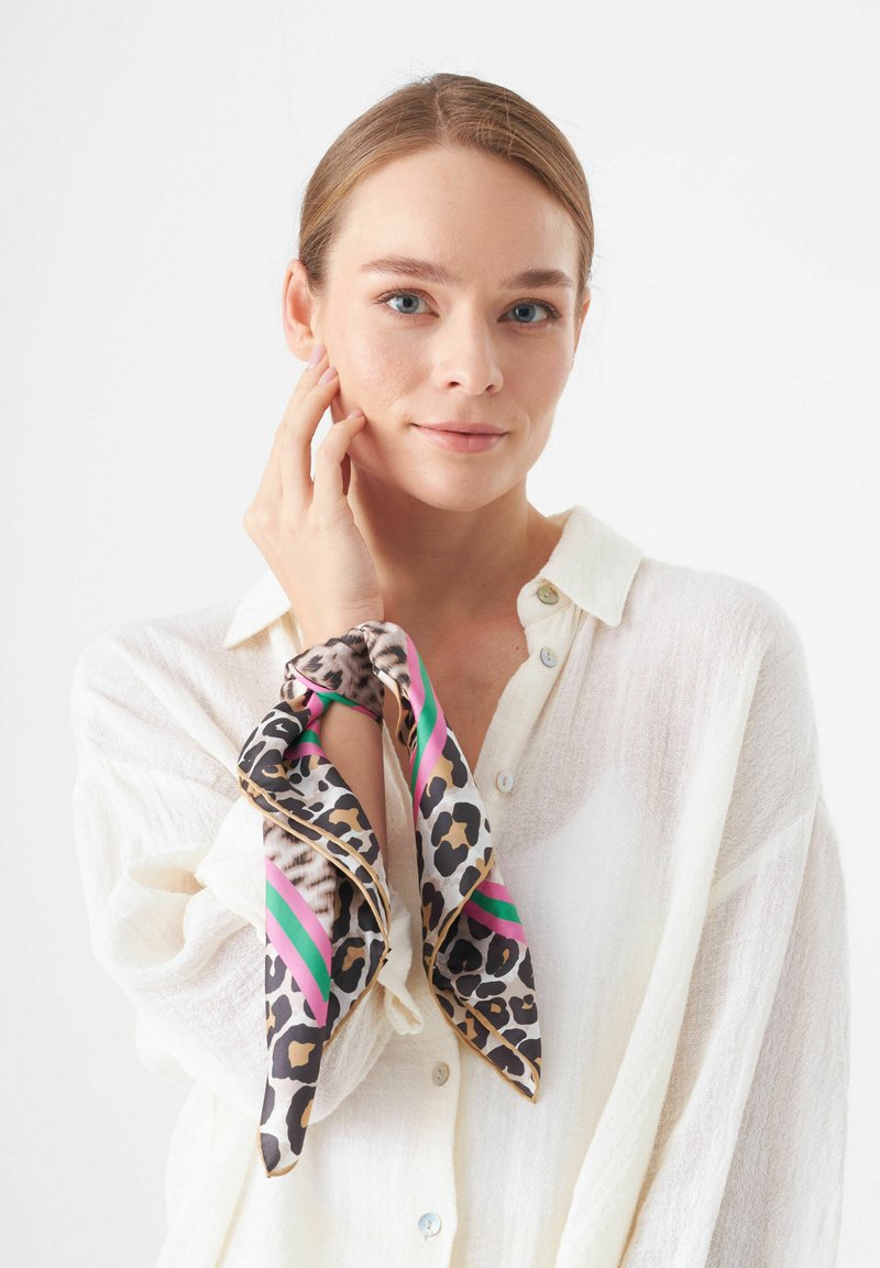 Leopardenprint zijden sjaal met roze en groene strepen, geknoopt om de pols. Model draagt een witte, lichte knoopshirt.