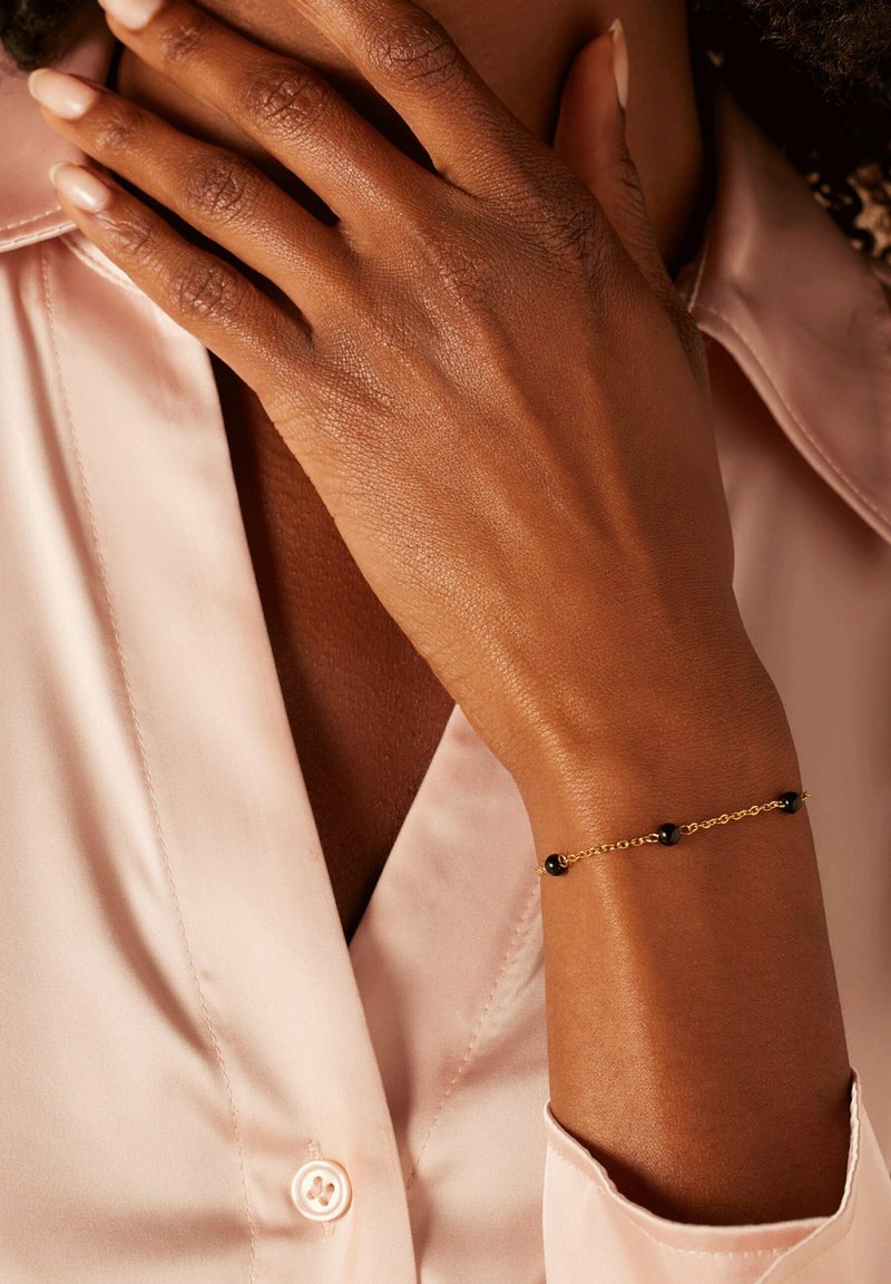 Goldene Kettenarmband mit schwarzen Perlen. Die Hand der Trägerin ruht auf einem hellrosa Satinhemd und präsentiert das Armband elegant.