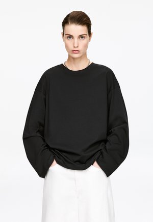 Young woman with slicked-back hair wearing a loose black long-sleeve top and white pants, standing with hands in pockets against white background.