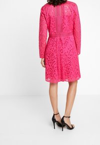 Vestido de encaje rosa con recortes geométricos, mangas largas y cintura ajustada. Combinado con sandalias de tacón alto negras.