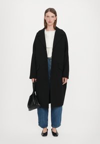 Lång svart ullkappa med stora slag, knappar framtill, kombinerad med en vit blus och blå vida jeans. Svart handväska som accent.