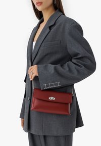 Rdeča usnjena torbica za ramo s srebrnim zapiranjem in verižnim ročajem, razstavljena ob sivi blazer s subtelnimi šivi.