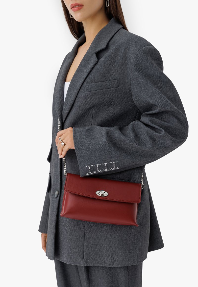 Rdeča usnjena torbica za ramo s srebrnim zapiranjem in verižnim ročajem, razstavljena ob sivi blazer s subtelnimi šivi.