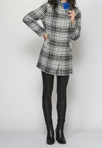 Kabát s károvaným vzorem v černé a bílé barvě, s dlouhými rukávy a kapsami, v kombinaci s černými koženými legínami a kotníkovými botami na podpatku.