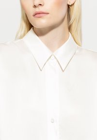 Weiße Seidenbluse mit einem klaren Kragen und einer Knopfleiste. Der Stoff hat eine glatte Textur mit einem dezenten Glanz.