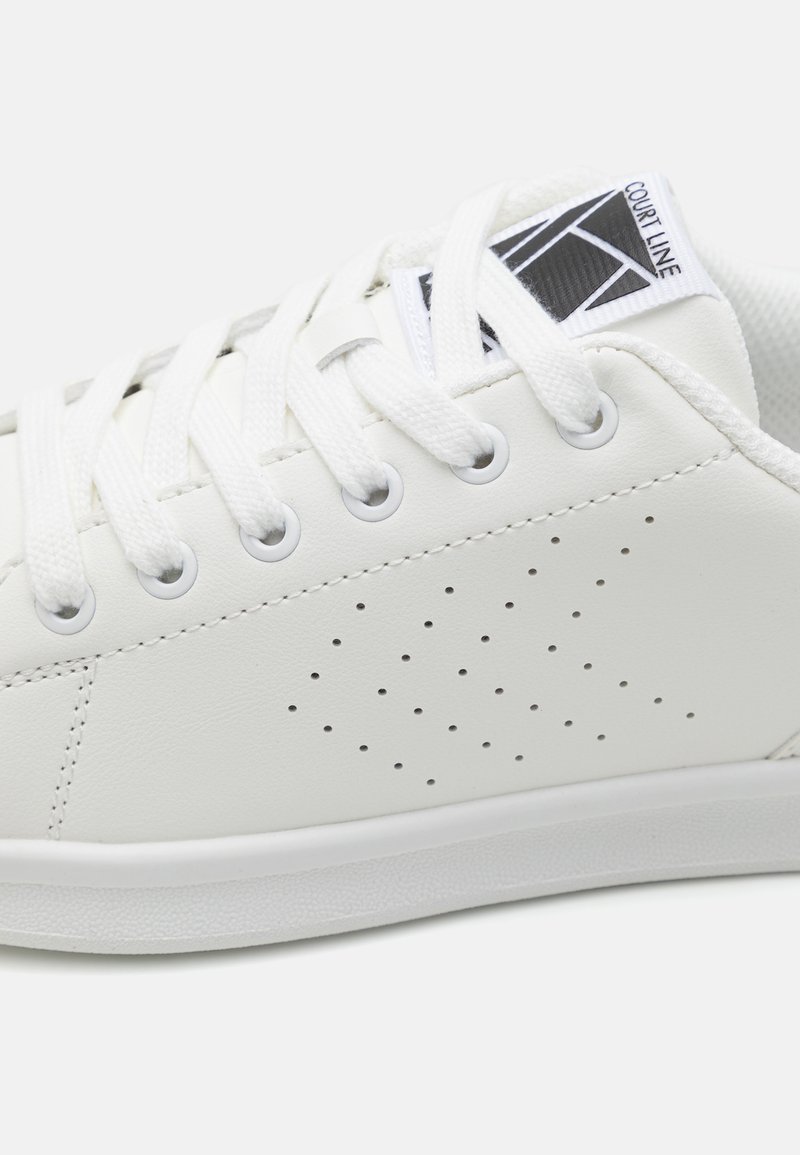 Zapatilla sintética blanca con detalles perforados, cordones redondos y suela texturizada. Etiqueta de logo en la lengüeta, diseño elegante.