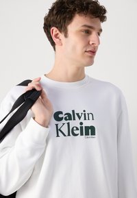 Biely sveter s zeleným textom "Calvin Klein", s vrúbkovými manžetami a okrúhlym výstrihom. Model drží čiernu tašku so popruhom. Jednoduchý dizajn, neformálny štýl.