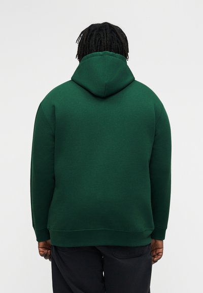 Oseba s kratkimi dreadlocksi, videna od zadaj, oblečena v temno zeleno kapuco in črne hlače na enobarvnem ozadju.