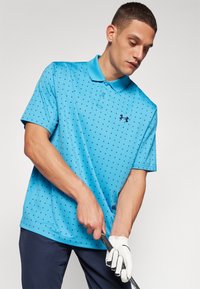 Cămașă polo albastră cu guler, având un model de puncte mici negre. Logo-ul Under Armour în negru pe pieptul stâng. Modelul poartă o mănușă de golf albă.