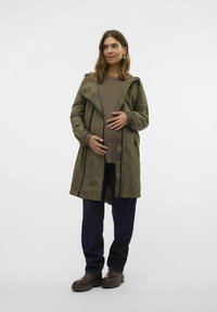 Giacca impermeabile verde oliva con zip frontale e cappuccio regolabile, indossata sopra un top marrone a coste e pantaloni blu scuro, abbinata a stivali marroni.