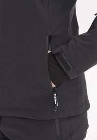 Svart mjukskaljacka med dragkedjor i sidfickorna, kardborrearmband och slät yta. Har en hög krage och figursydd design.