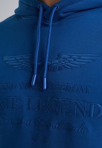 Blue hoodie with embossed text and wing design. Features drawstrings and a soft texture. Details include "FIND YOUR FREEDOM" and "1958" embossed.