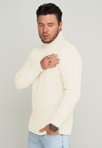 Cremefarbener, gestrickter Rollkragenpullover mit gerippten Bündchen und Saum, kombiniert mit hellblauer Jeans. Accessoires sind Ringe und eine Uhr.