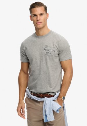 Camiseta gris de algodón de manga corta con un logo azul de Superdry & Co en el pecho, combinada con pantalones beige y una camisa a rayas azules atada a la cintura.