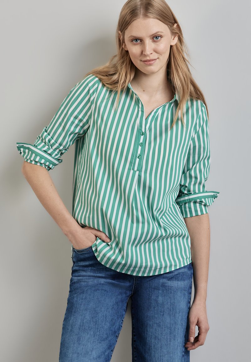 Blusa a righe verdi e bianche con una pattina con bottoni, maniche arrotolate e una vestibilità rilassata, abbinata a jeans blu. Tessuto morbido.