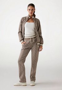 Brązowy wzorzysty dres z kurtką na zamek i spodniami ze ściągaczem, z białymi akcentami i dopasowanym topem na ramiączkach pod spodem. Białe sportowe buty.