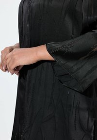 Schwarzes Kimono mit einem strukturierten Muster und weiten Ärmeln. Der Stoff wirkt glatt und leicht glänzend und weist subtile Designdetails auf.