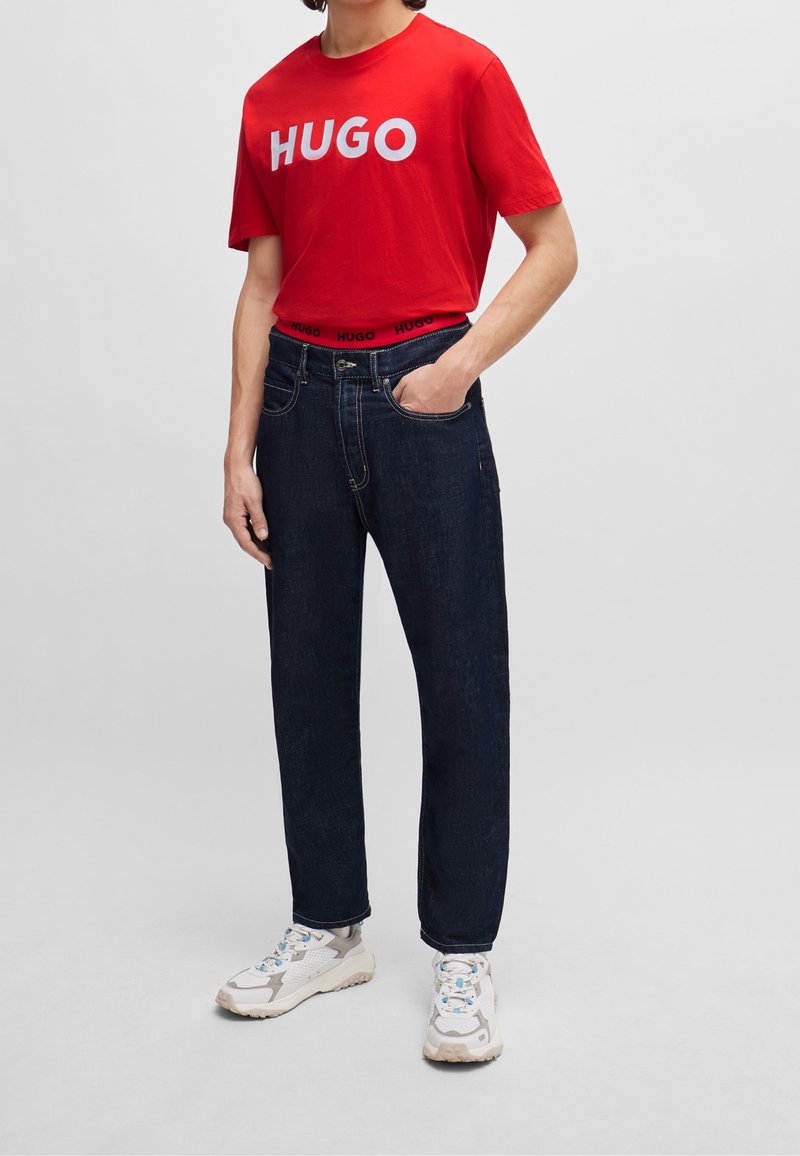 T-shirt en coton rouge avec le logo "HUGO", jean en denim bleu foncé et baskets grises avec des accents beige. Coupe décontractée avec des coutures détaillées.
